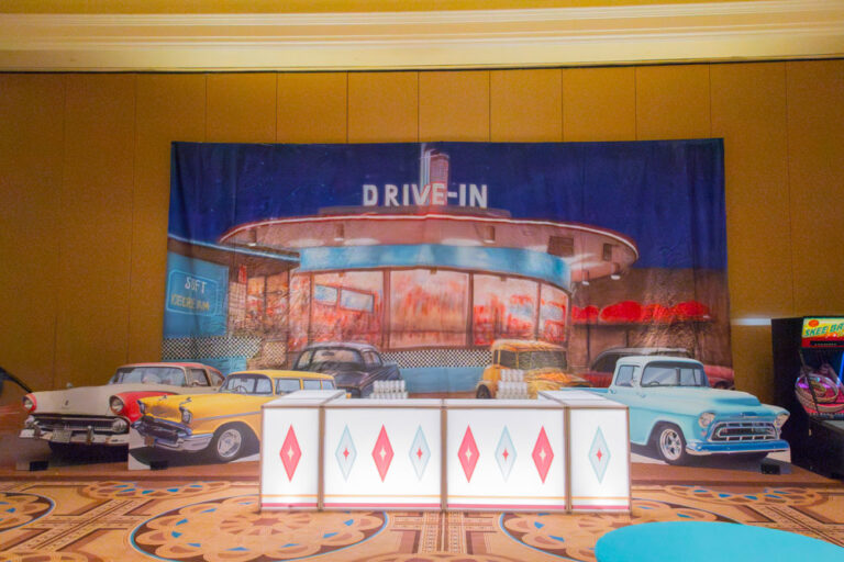 Retro drive-in diner backdrop with vintage car cutouts and diamond-accented bar at themed event