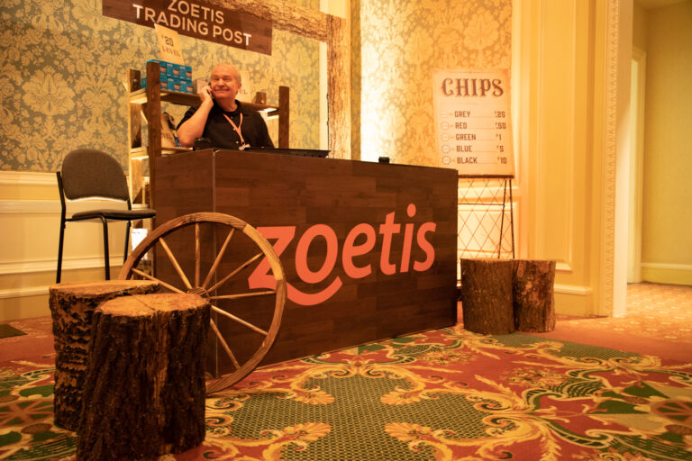 Western-themed Zoetis trading post booth with wood accents, wagon wheel decor, and a chip value sign, staffed by a smiling man on the phone.