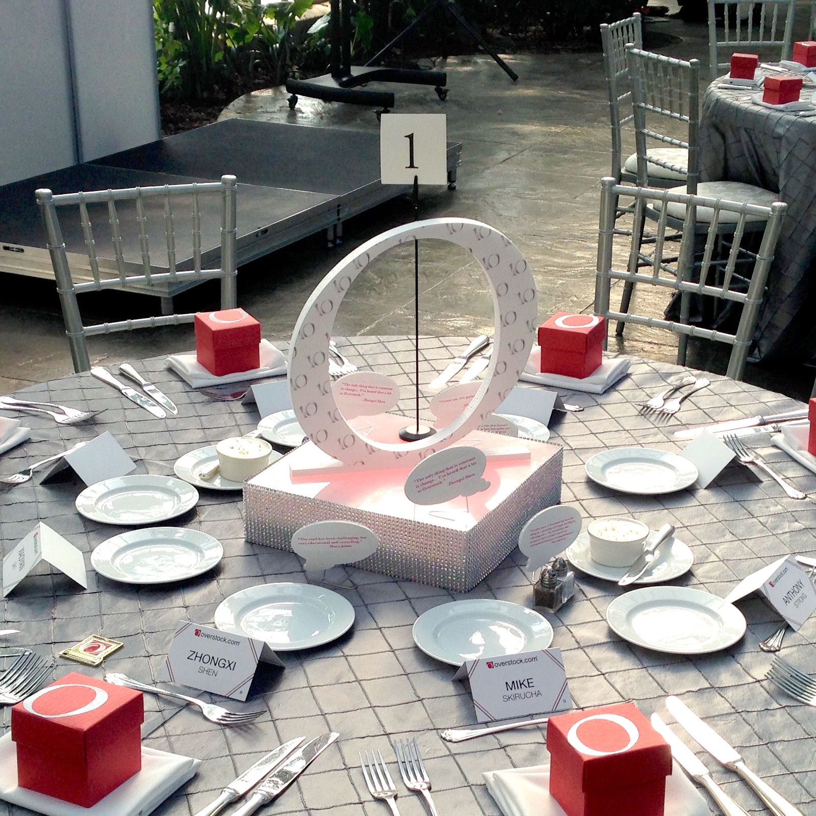 Elegant event table with gray linens, silver chairs, red gift boxes, and a custom Overstock O centerpiece.