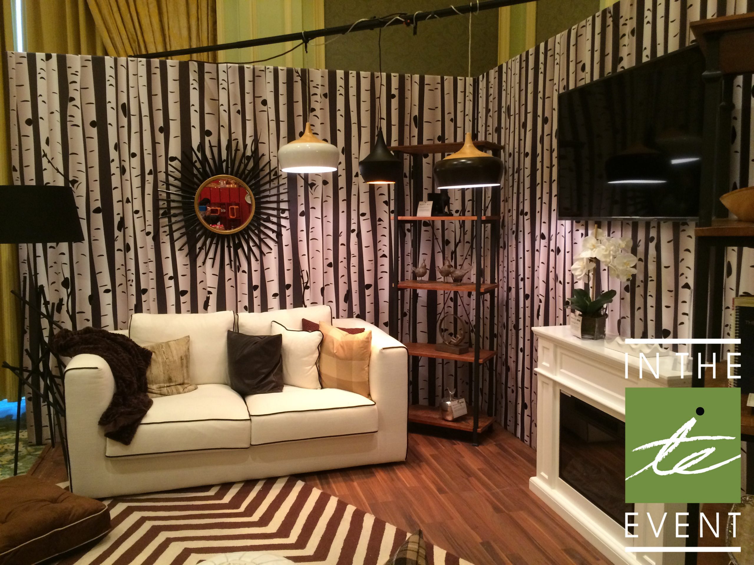 Styled Overstock living room activation with birch tree backdrop, white sofa, modern lighting, fireplace, and shelving accents.