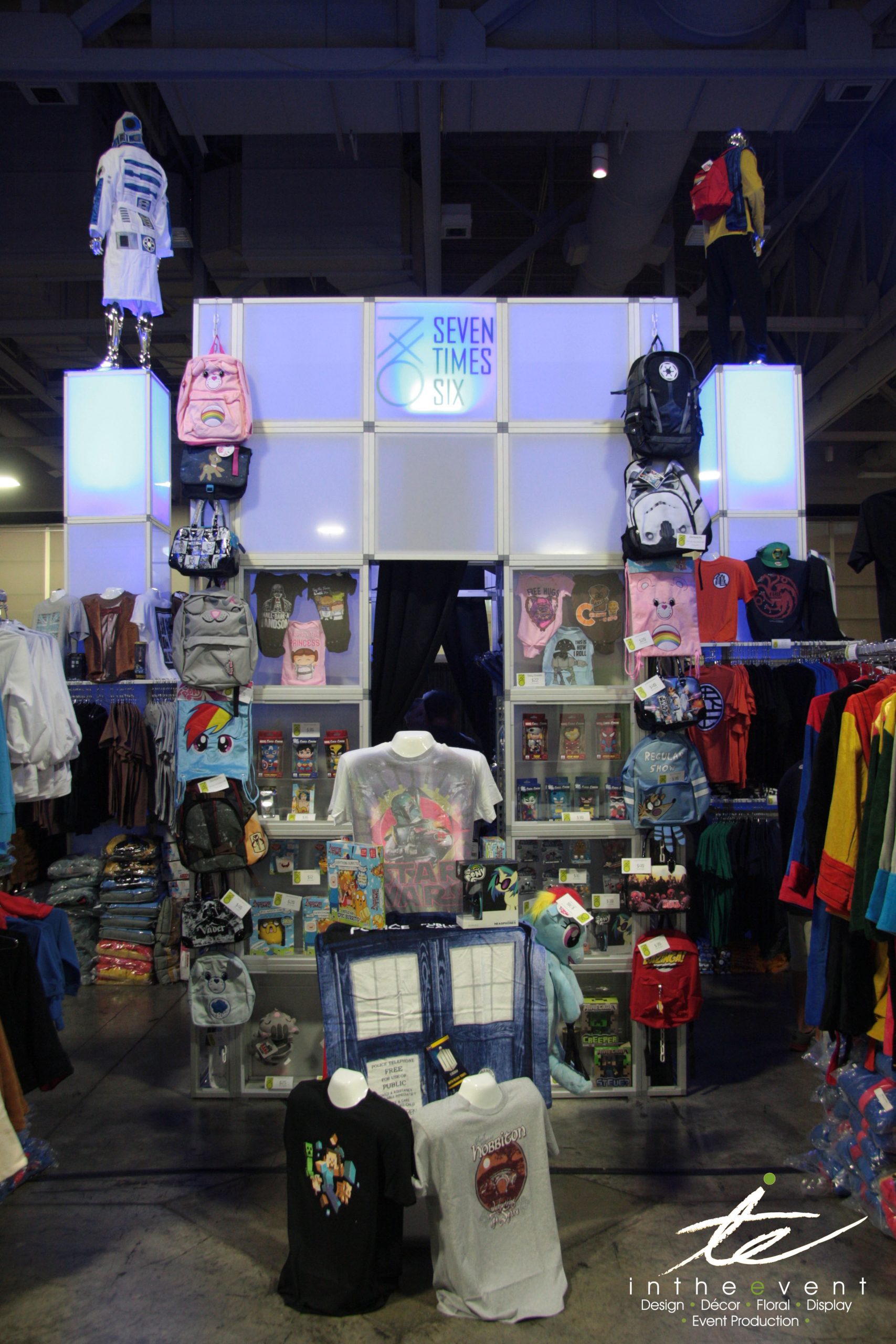 Trade show booth for Seven Times Six featuring modular illuminated walls and towers by In The Event, showcasing apparel, backpacks, and pop culture merchandise.