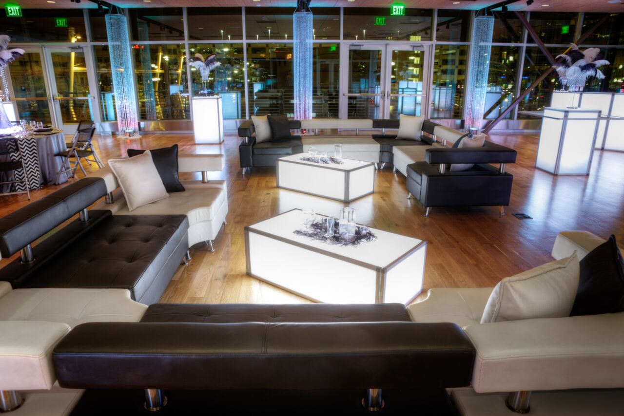 Stylish event lounge with modular black and white sofas, glowing LED cocktail tables, and decorative uplighting.