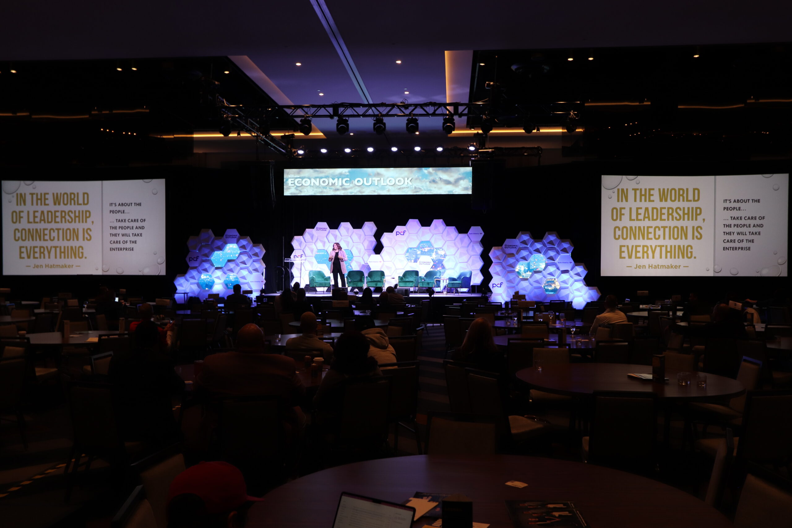 Conference stage setup with hexagonal LED backdrop, projection screens, and keynote speaker addressing audience.