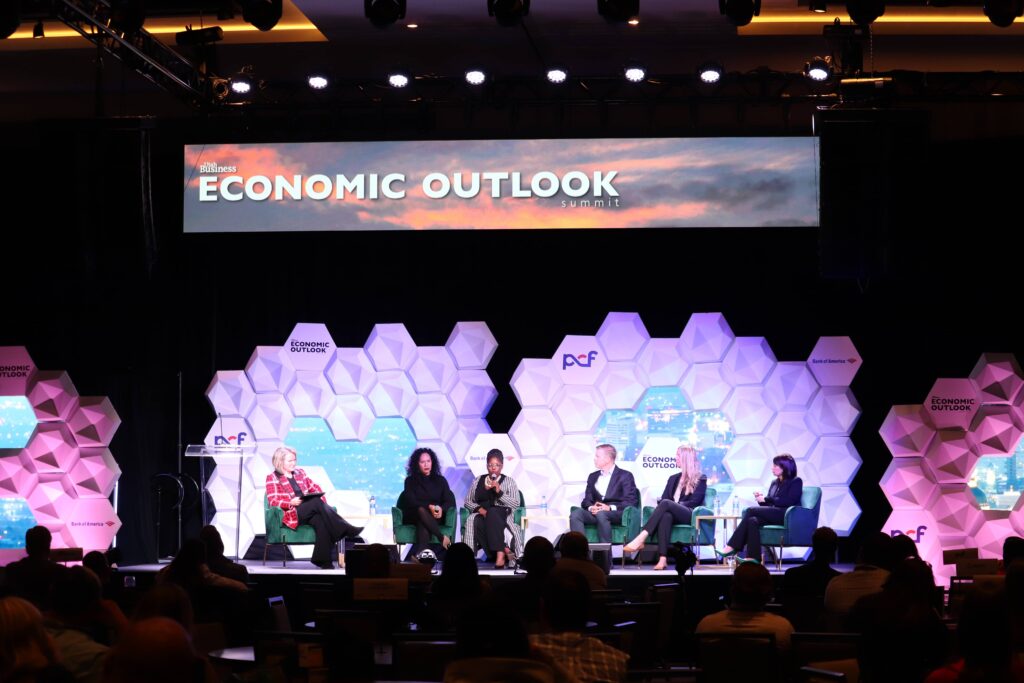 Conference stage with hexagonal LED panel backdrop, large projection screens displaying a leadership quote, and a presenter speaking to an audience at the Economic Outlook event.