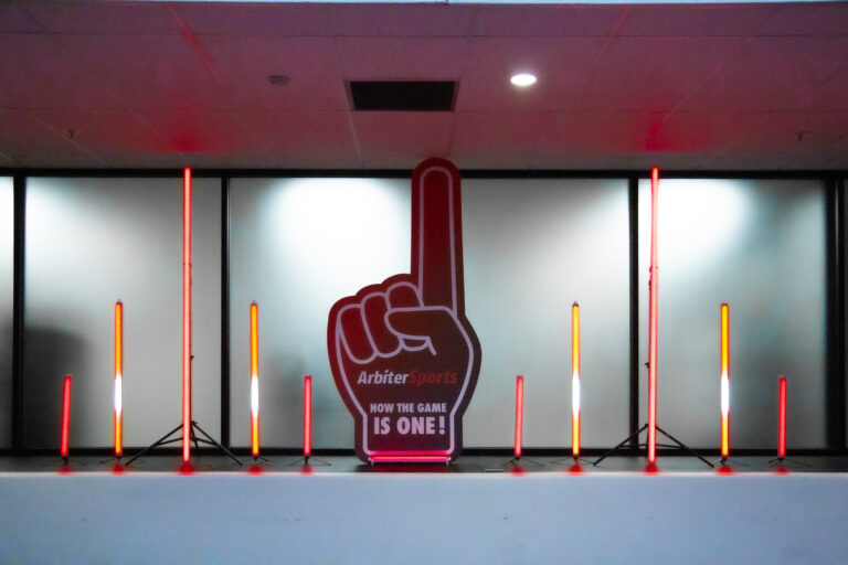 Oversized foam finger cutout with the Arbiter Sports logo reading “How the Game Is One!” surrounded by vertical red and white LED light columns.