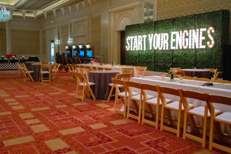 Wide-angle view of an event space featuring a large illuminated "START YOUR ENGINES" marquee wall, racing arcade games, and checkered flag decor.