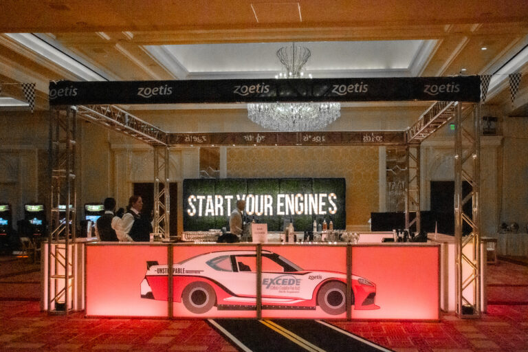 Illuminated race-themed bar with a printed sports car wrap, overhead Zoetis trusses, and a glowing “Start Your Engines” sign in the background.