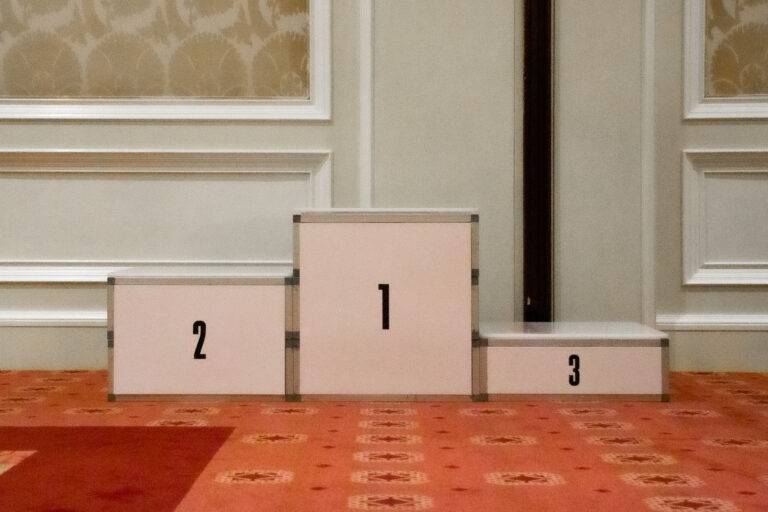 A white three-tier winner’s podium with black numbers 1, 2, and 3, set against an elegant ballroom wall.