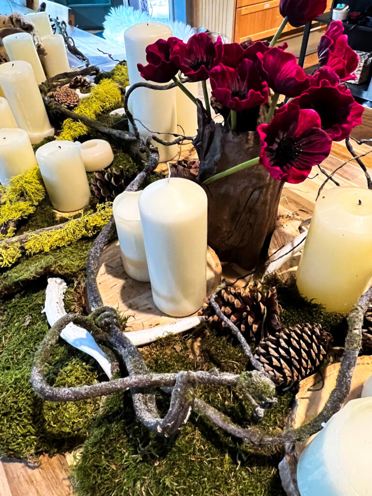 Close-up of a detailed woodland table arrangement featuring moss, antlers, white candles, pinecones, and a vase of dark red flowers.