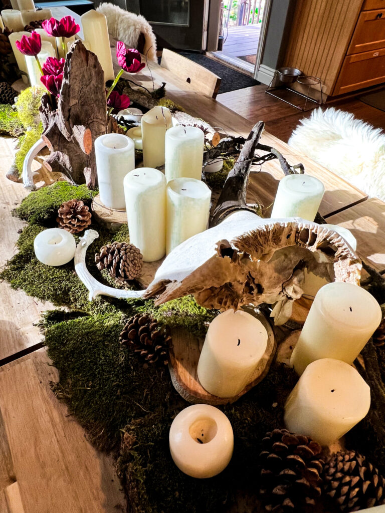 Long wooden table decorated with moss, pinecones, antlers, white pillar and votive candles, dark red flowers, and a skull.