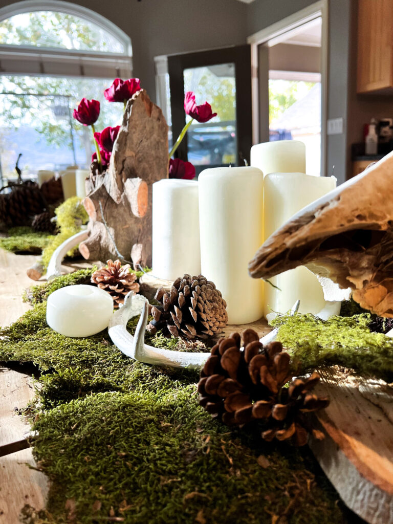 Close-up of an indoor table centerpiece featuring moss, pinecones, antlers, white candles, and dark red flowers.