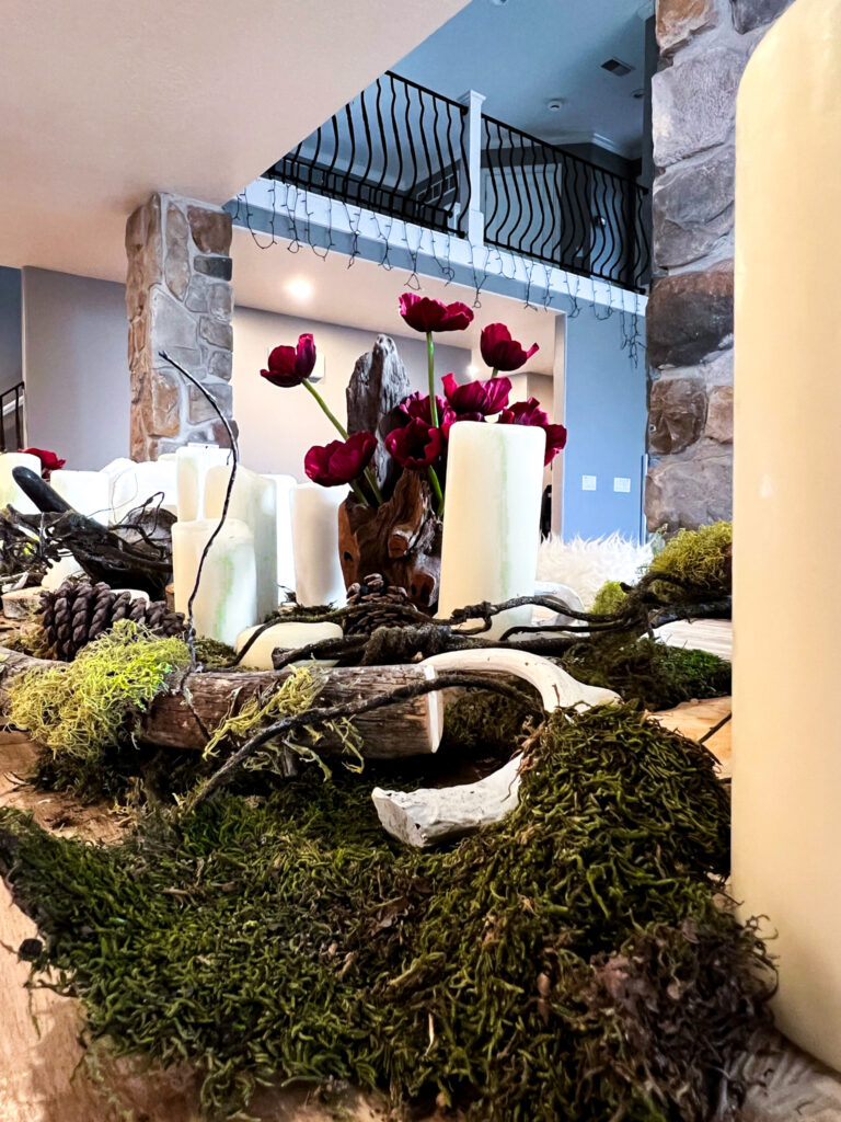 Detailed view of a woodland-themed table centerpiece with moss, branches, dark red flowers, and white candles.