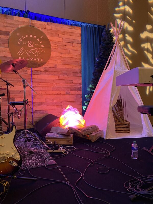 Rustic stage setup with wooden backdrop, faux campfire, teepee, trees, and musical instruments at Celebrate & Elevate event