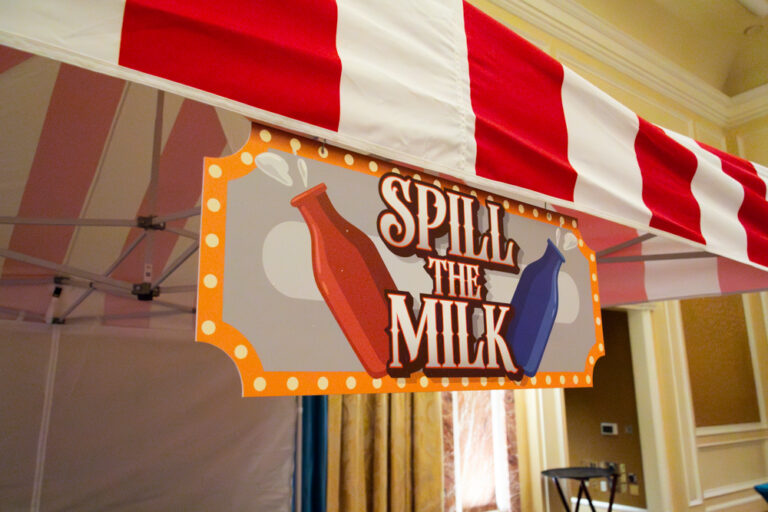 Carnival game sign reading "Spill the Milk" under red and white striped canopy