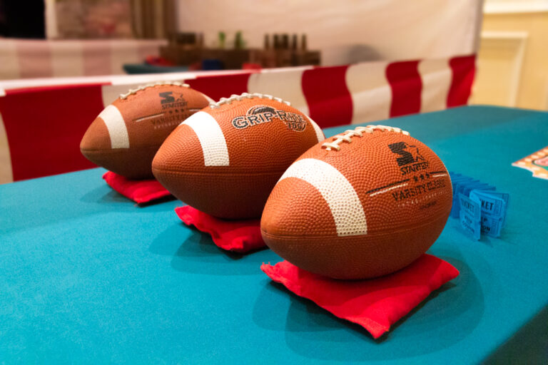 Three footballs on red cloth pads arranged on a teal table at a carnival-themed game booth with a red and white striped backdrop.