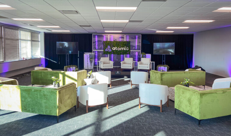 Corporate event stage with Atomic branding, greenery backdrop, and modern lounge seating in front.