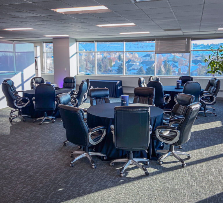 Round tables with black linens surrounded by leather rolling office chairs in a sunlit event space.