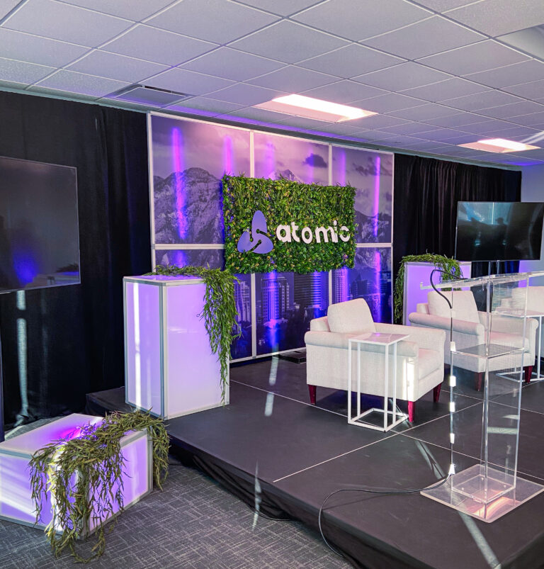 Event stage with two cream armchairs, greenery-wrapped Atomic logo, and purple uplighting in front of a cityscape and mountain backdrop.