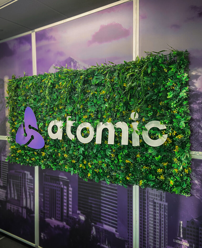 Atomic logo mounted on a rectangular green wall with lush foliage and small yellow flowers, set against a purple cityscape backdrop.