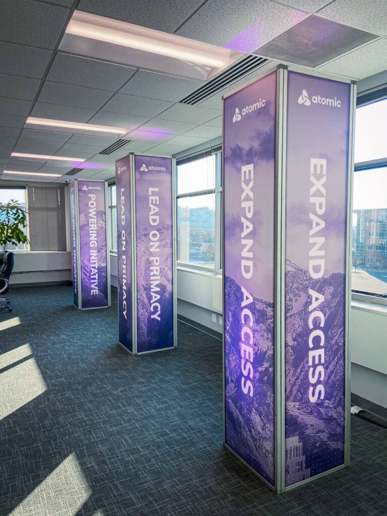 Branded purple pillar displays with Atomic logo and value statements like “EXPAND ACCESS” and “LEAD ON PRIMACY.”