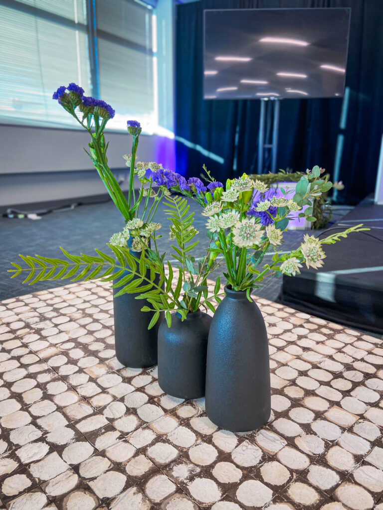 Three matte black vases with greenery and purple flowers on a patterned table in front of Atomic stage setup. Custom floral arrangement for event reception table