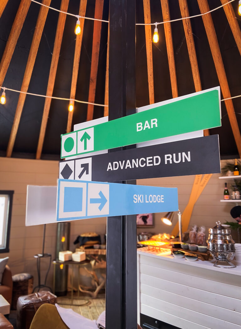 Directional ski-themed sign inside a cozy alpine lounge with leather chairs and string lights.