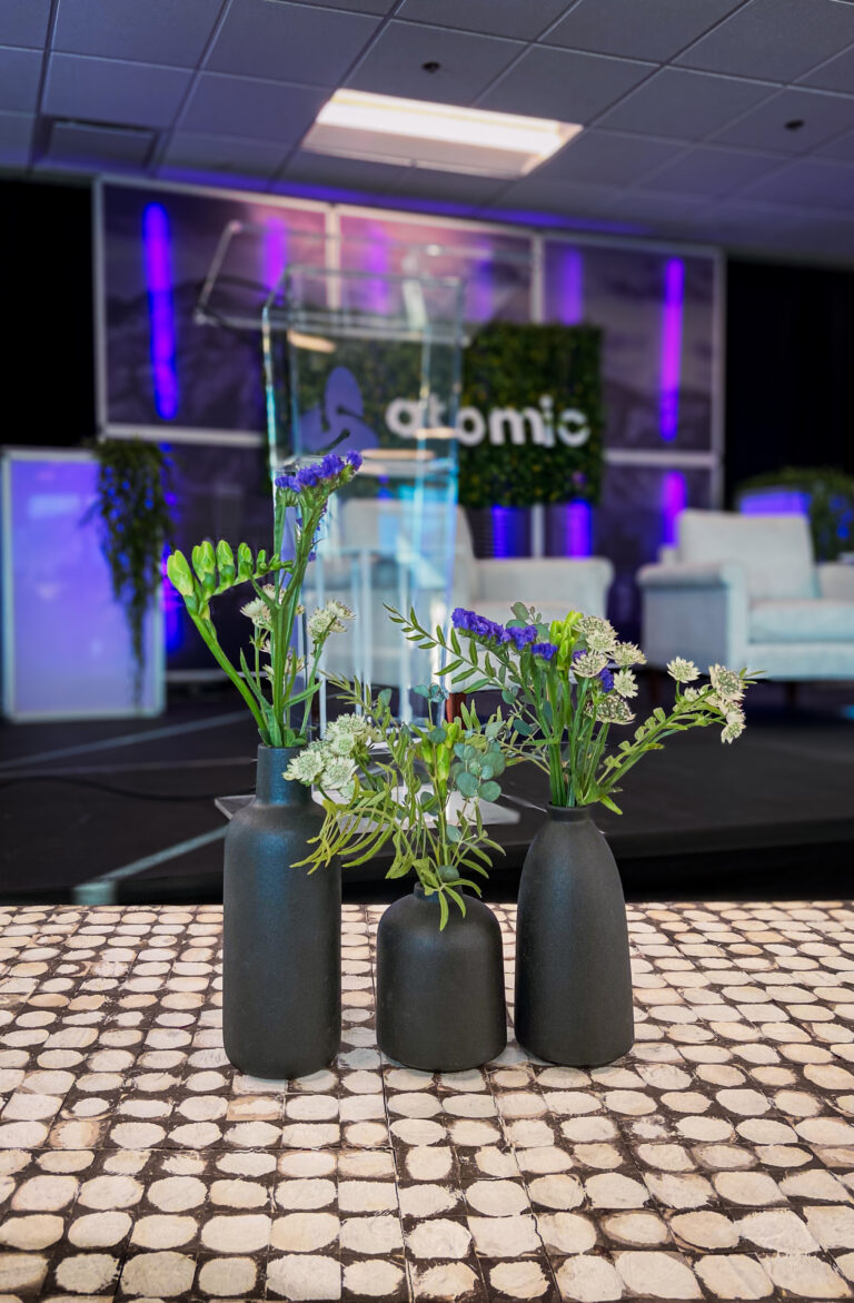 Three matte black vases with greenery and purple flowers on a patterned table in front of Atomic stage setup.