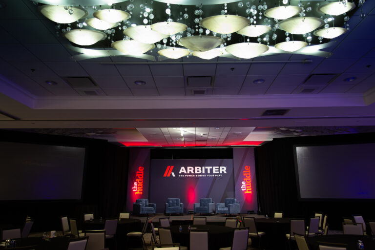 Stage setup for a branded panel event with four blue chairs and bold Arbiter and “The Huddle” signage.
