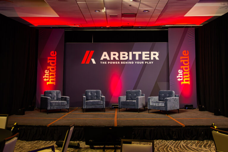 Stage setup for a branded panel event with four blue chairs and bold Arbiter and “The Huddle” signage.