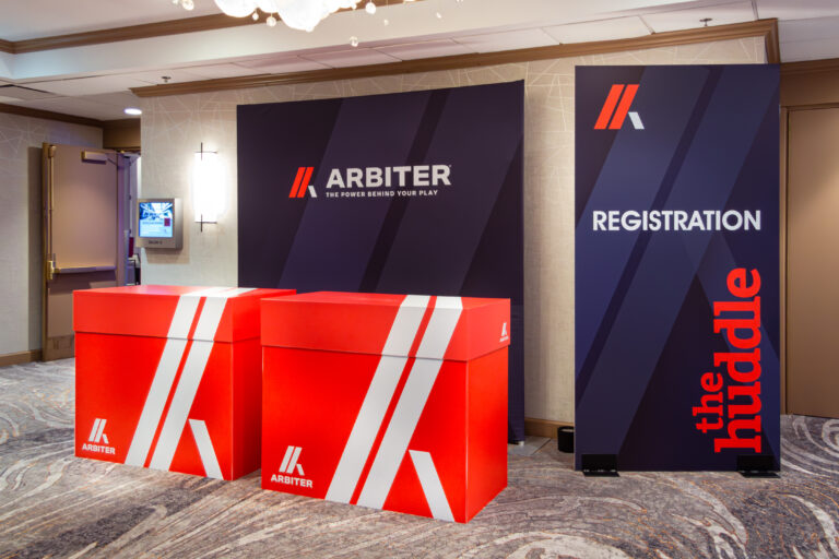 Branded event registration area with bold red and navy signage for Arbiter and “The Huddle.”