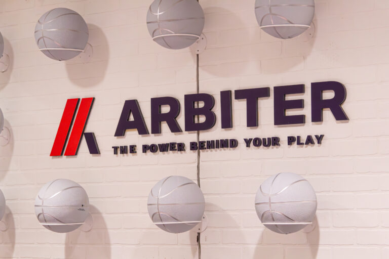 Close-up of Arbiter logo and tagline on a white brick wall with mounted silver basketballs.