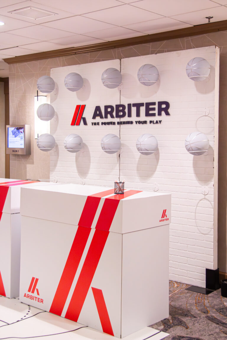 Branded registration counters with a basketball court floor graphic and a backdrop of silver basketballs and Arbiter signage.