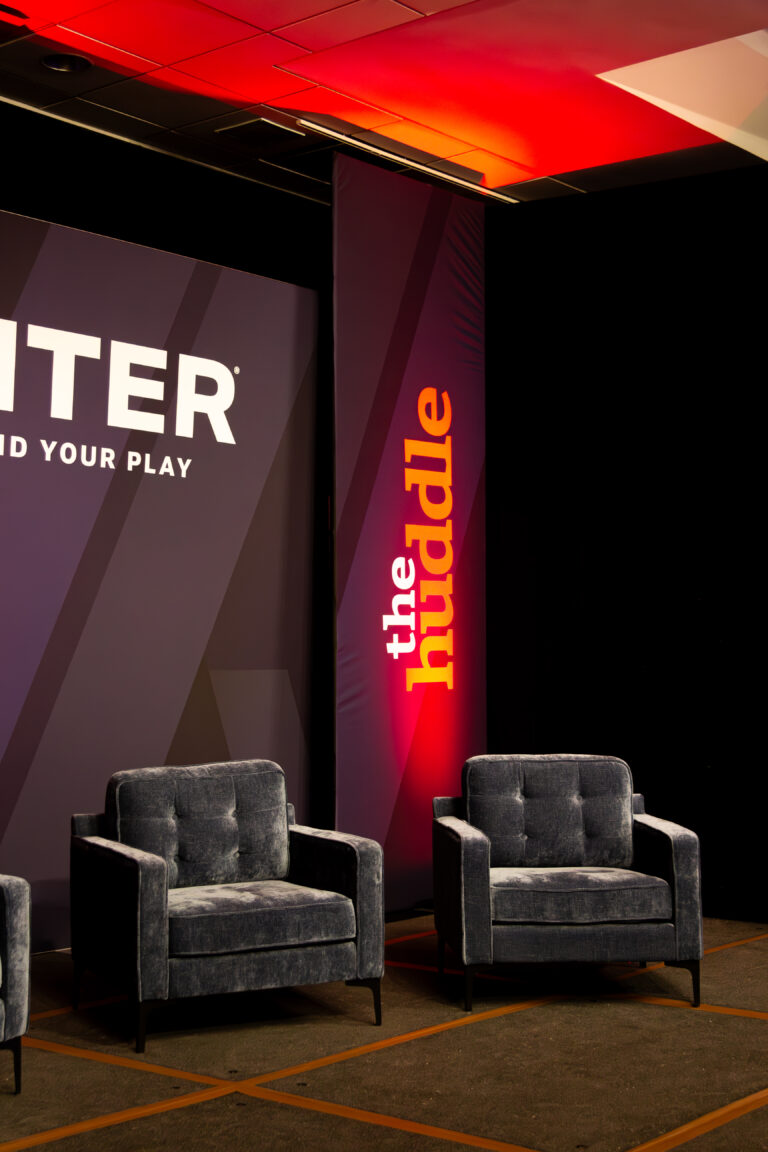 Close-up of two modern blue velvet chairs on stage with “The Huddle” branding and dramatic red uplighting.