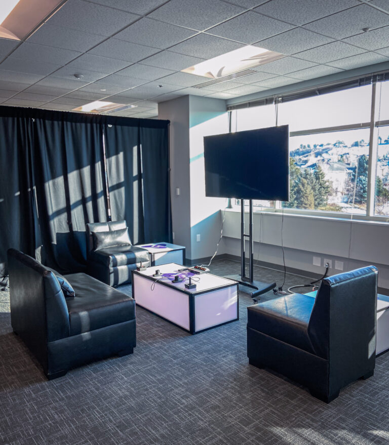 Black lounge seating around a light-up gaming table with TV setup in front of large windows.