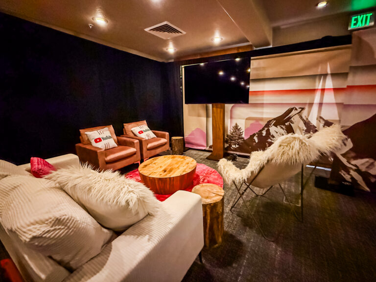 A warm lounge corner with a cream sofa, leather chairs, fluffy throws, rustic wood tables, and YouTube-branded pillows set against a mountain-themed backdrop.