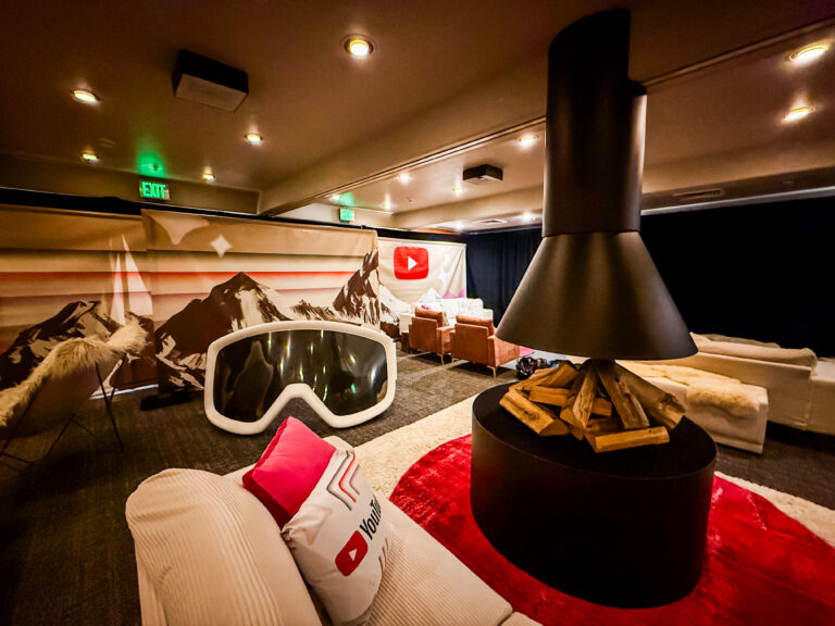 A cozy branded lounge featuring a central faux fireplace, oversized ski goggle decor, mountain graphics, and YouTube-themed pillows and signage. Contemporary lounge setup including sofa and table, part of our event design services.