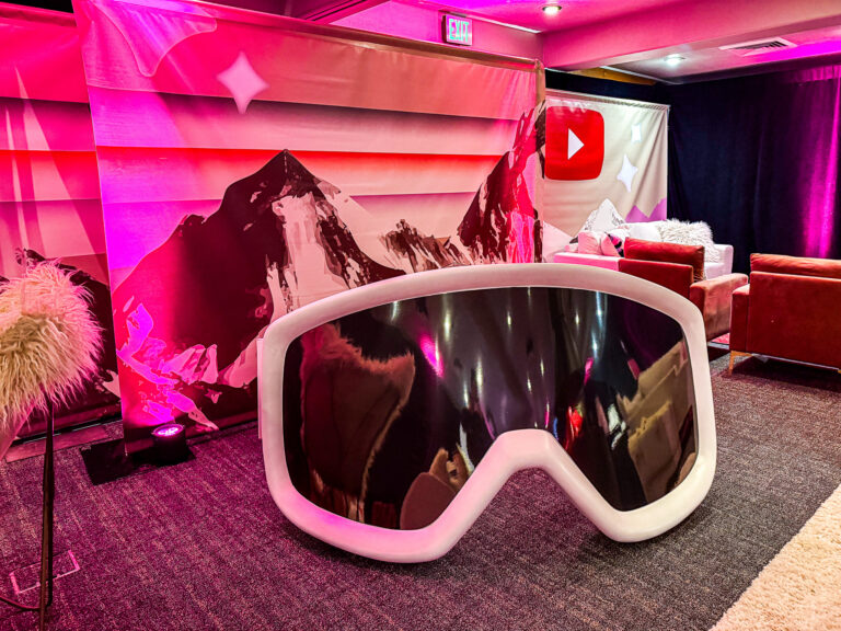 A colorful lounge space with oversized ski goggles, red velvet seating, faux fur accents, and a custom mountain backdrop featuring the YouTube logo.