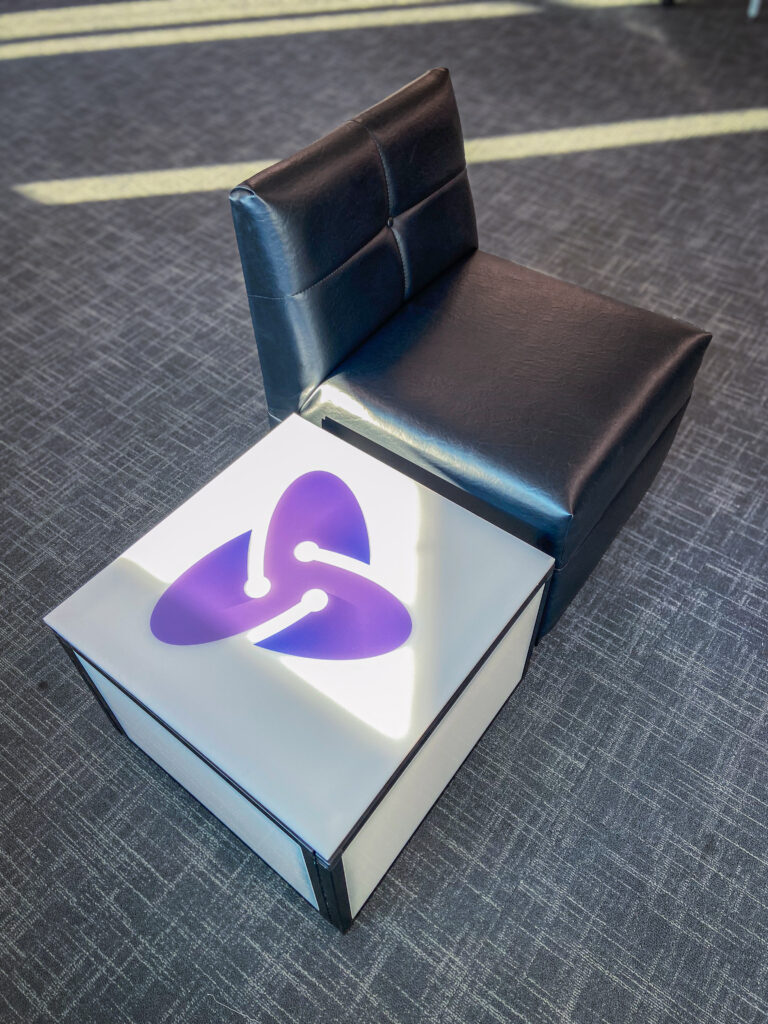 Black modular lounge chair next to branded light-up cube with Atomic logo on carpeted flooring.