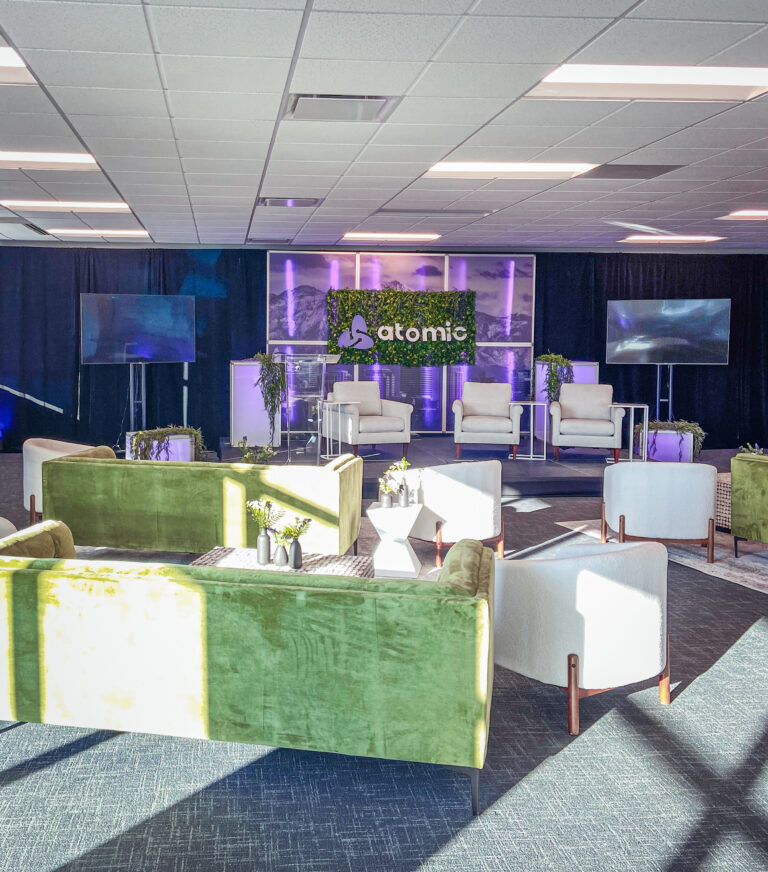 Corporate event stage with Atomic branding, greenery backdrop, and modern lounge seating in front.