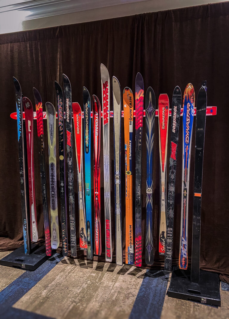 A decorative wall installation made from upright vintage skis in a range of colors and brands, arranged side-by-side in metal stands against a brown curtain backdrop.