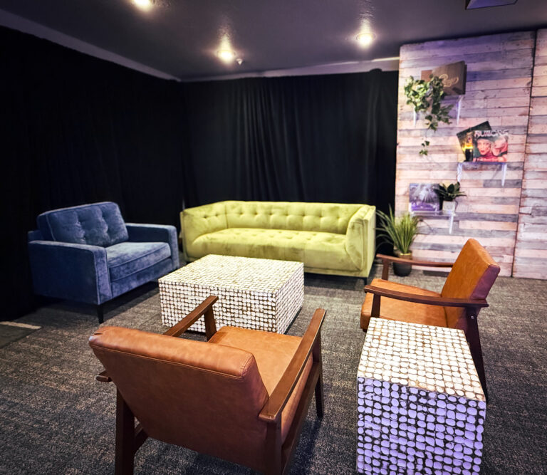 Eclectic lounge setup with a green velvet sofa, blue armchair, two leather chairs, and mosaic cube tables in front of a wood panel wall with vinyl records.