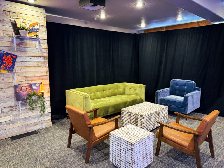 Eclectic lounge setup with a green velvet sofa, blue armchair, two leather chairs, and mosaic cube tables in front of a wood panel wall with vinyl records.