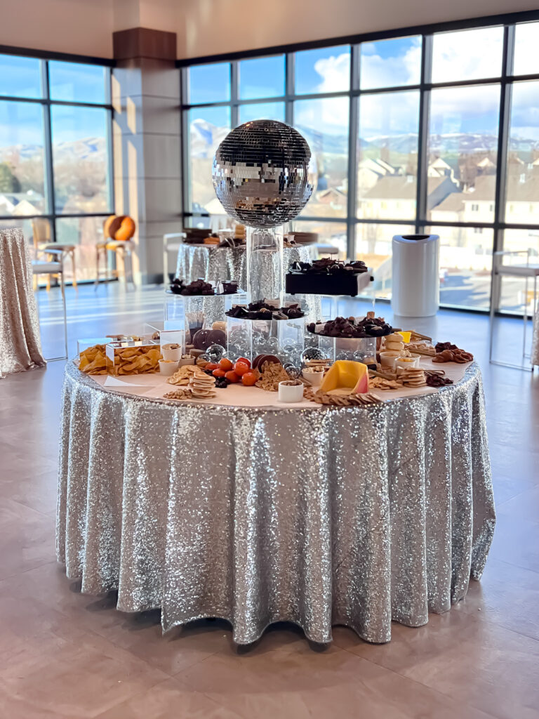 Grazing table with disco ball centerpiece, assorted snacks, and silver sequin tablecloth.