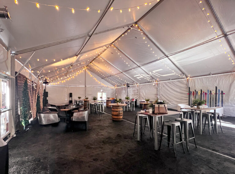 Wide view of a tented event lounge with barrel cocktail tables, high-top seating, metallic armchairs, ski-themed decor, and overhead string lights.