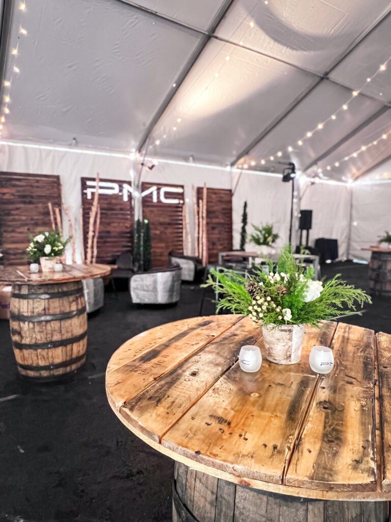 Rustic event lounge inside a tent with wooden barrel cocktail tables, greenery centerpieces, aluminum chairs, and branded wood-paneled backdrop.
