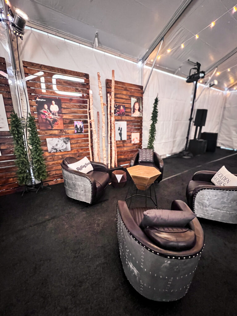 Cozy lounge vignette with four metallic armchairs, a geometric side table, branded pillows, and rustic wood paneling with event photos inside a tented space.