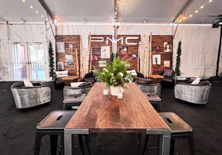 Tented lounge setup with industrial-style high tables, aluminum armchairs, rustic wood accent walls with photo displays, and winter greenery decor.