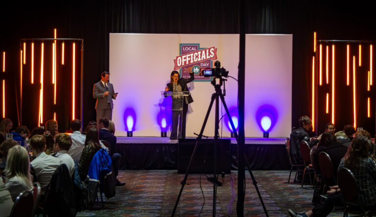 A wide view of a live event with two presenters on stage, vertical LED tube lighting, a white branded backdrop, and audience seating in the foreground.