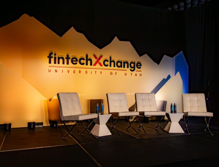 A modern panel stage setup for FintechXchange at the University of Utah, featuring a custom mountain-inspired backdrop, white leather chairs, and geometric side tables with water bottles.