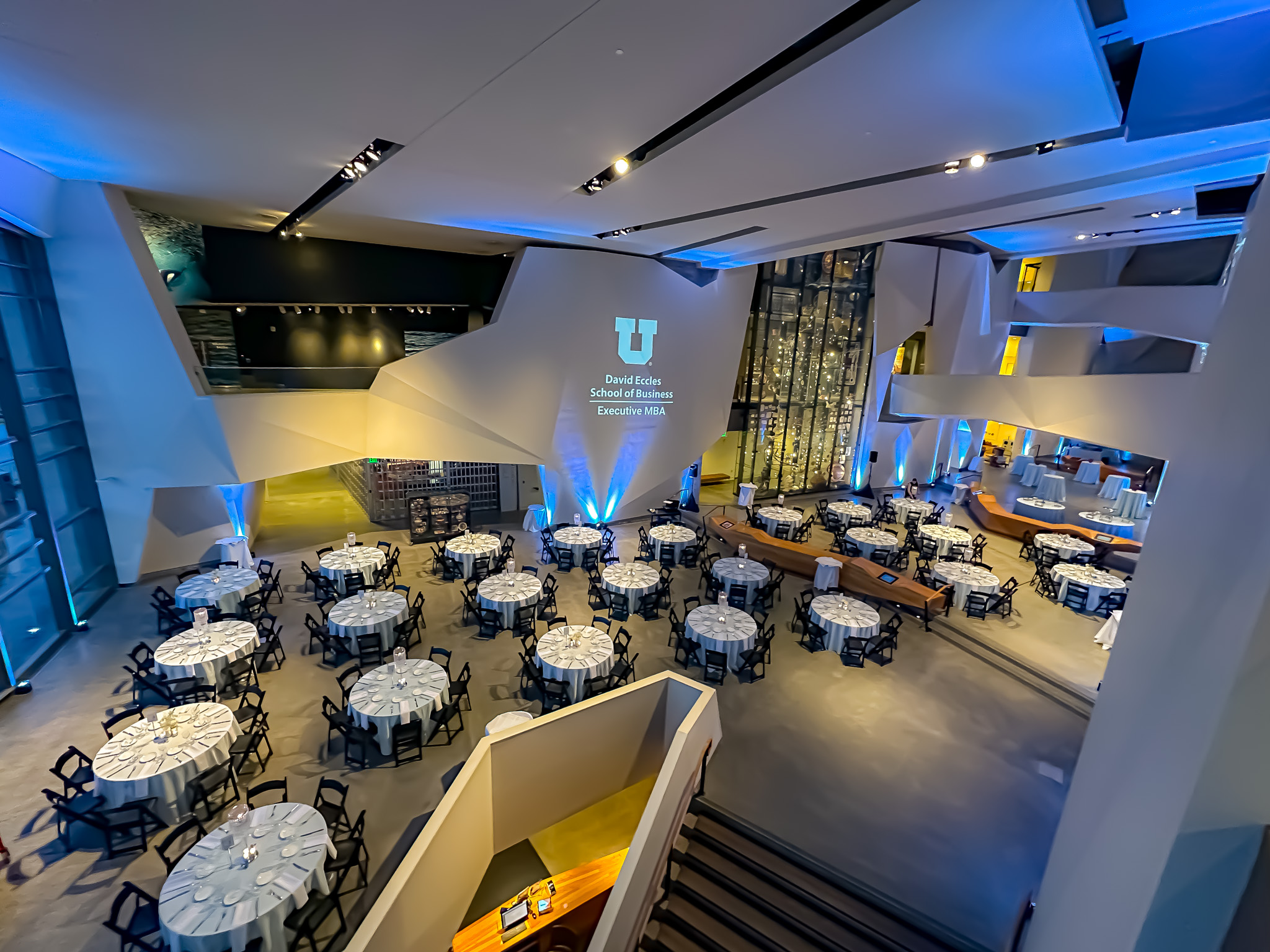 Elegant event setup with round tables covered in white linens, black chairs, and blue uplighting in a modern venue.